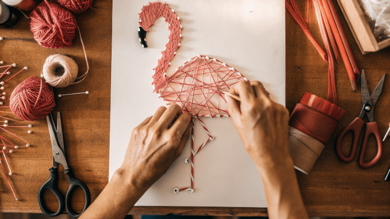 Adding Pink Thread to Flamingo Photograph of hands crafting a vibrant pink flamingo string art on a white board, surrounded by craft supplies, evoking creativity and DIY satisfaction.