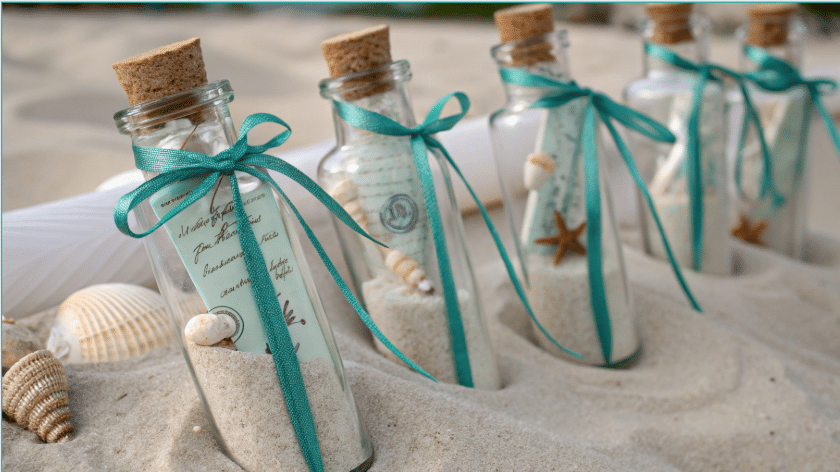 Mermaid Party Favors Photograph of small glass bottles filled with sand and rolled messages, adorned with teal ribbons and seashells, suggesting a beach or wedding theme.