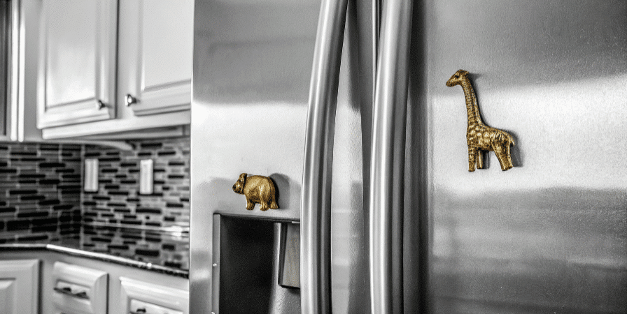 Photograph of two tiny golden animal-shaped magnets (, giraffe, elephant) adorning a sleek, modern stainless steel refrigerator in a clean, bright kitchen setting.