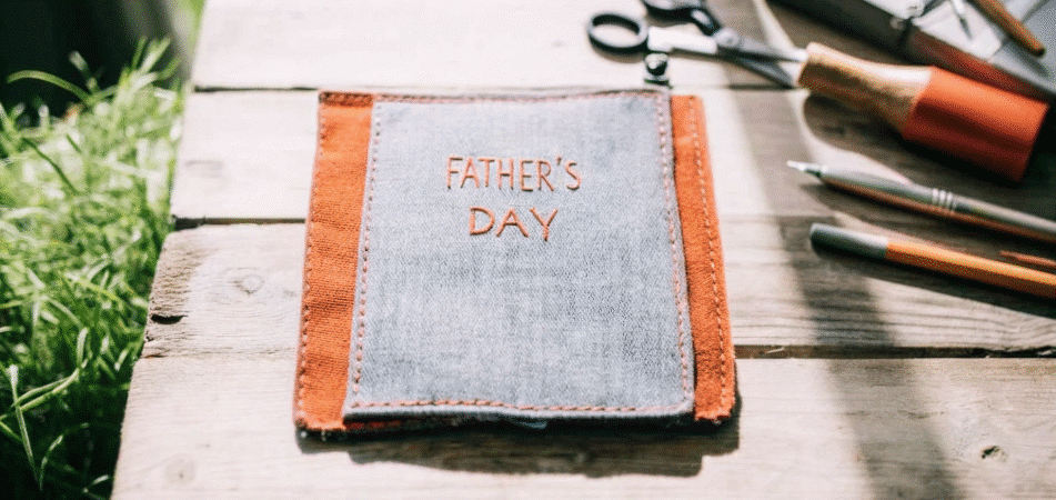 Photograph of a handmade denim and orange fabric pouch with 'Father's Day' embroidery, surrounded by crafting tools on a rustic wooden surface, evoking warmth and thoughtfulness.
