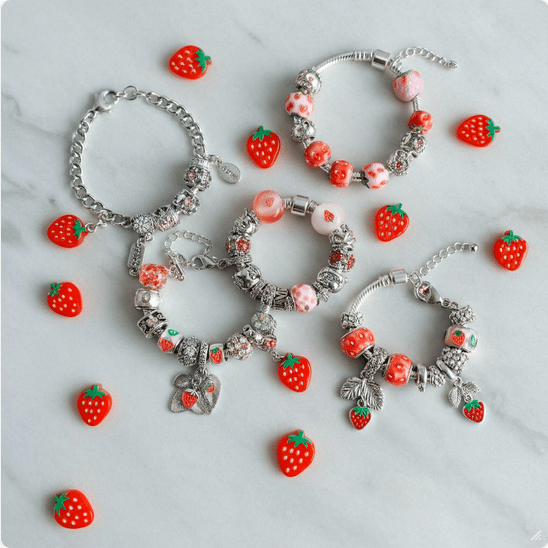 Strawberry Bracelet Flat Lay Photograph of multiple silver charm bracelets adorned with red and pink strawberry-themed beads and charms, displayed on a pristine white marble surface, evoking sweetness and personalization.