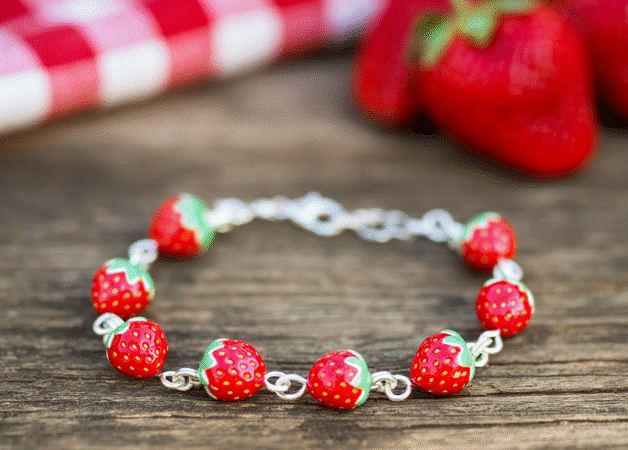 Photograph of a charming strawberry-themed bracelet on a rustic wooden table, with fresh strawberries and a red gingham picnic blanket blurred in the background, evoking summer and sweetness.