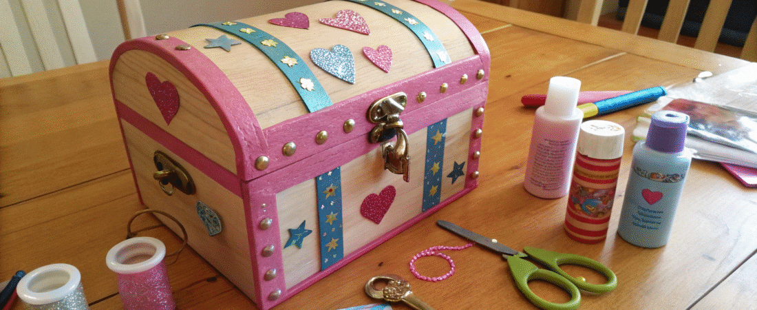 A photo featuring a creative crafting scene with a focus on a detailed and colorful craft project. The main object is a wooden chest-like container, adorned with various embellishments such as heart-shaped cutouts, glittery details, and other decorative elements.