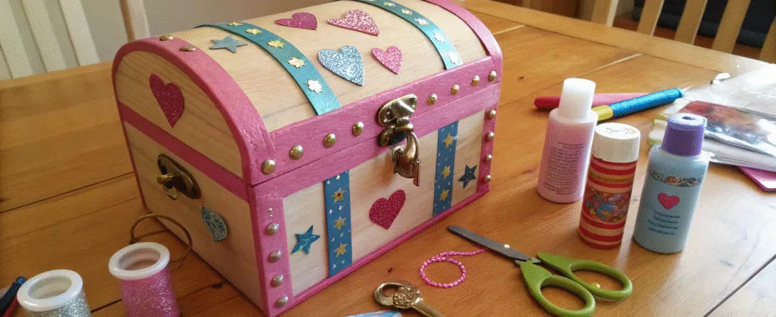 A photo featuring a creative crafting scene with a focus on a detailed and colorful craft project. The main object is a wooden chest-like container, adorned with various embellishments such as heart-shaped cutouts, glittery details, and other decorative elements.
