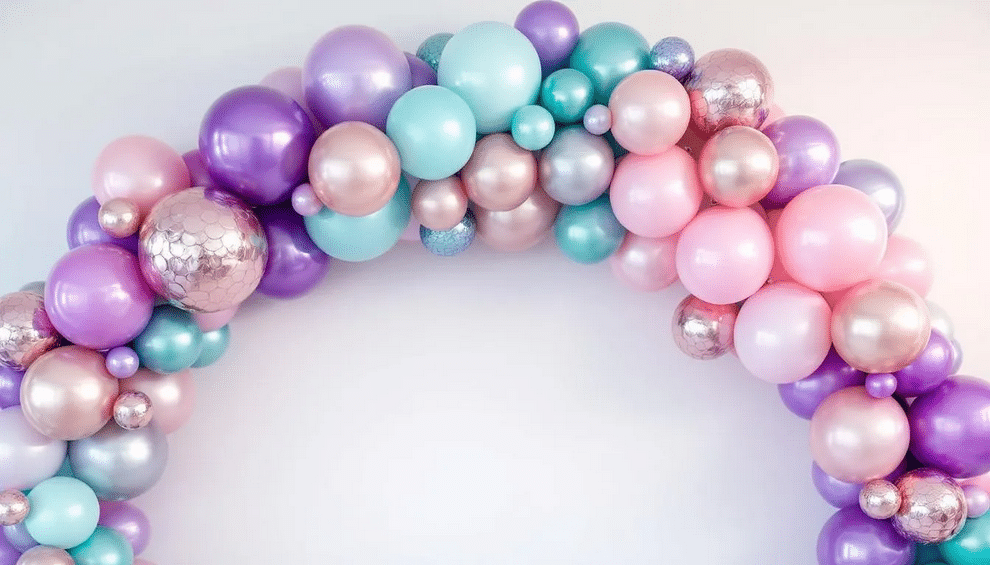 A vibrant balloon arch features shades of purple, teal, and soft pink, reminiscent of the sea, with some balloons showcasing metallic and pearl finishes for a shimmering effect. Bubble balloons add texture, creating a playful atmosphere perfect for a mermaid-themed party.