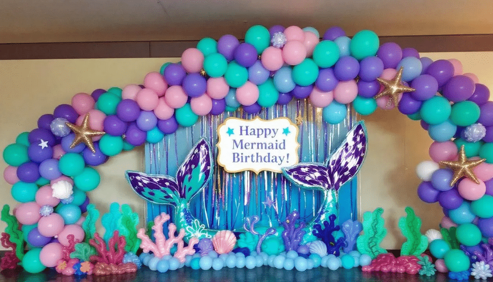 The image showcases a vibrant mermaid balloon feature, including a large mermaid tail balloon at one end of a colorful balloon garland. The arrangement features shades of purple, pink, and teal, along with whimsical sea-themed balloons, creating a magical atmosphere perfect for a mermaid party.