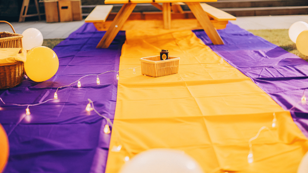 An outdoor pop up birthday party setup features a colorful picnic table adorned with a vibrant tablecloth, surrounded by playful decorations like balloons and string lights, creating a fun and inviting space for guests to celebrate. The atmosphere is enhanced by the presence of picnic blankets and baskets, making it a memorable experience for kids and adults alike.
