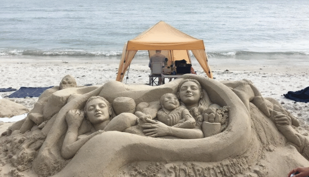 The image shows a beautifully crafted sand sculpture, meticulously detailed and standing proudly on a sandy beach. Surrounding the sculpture are tools like shovels and buckets, indicating the sand sculpting process, while a few spectators admire the artwork under the shade of a tent, ensuring its preservation from the elements.