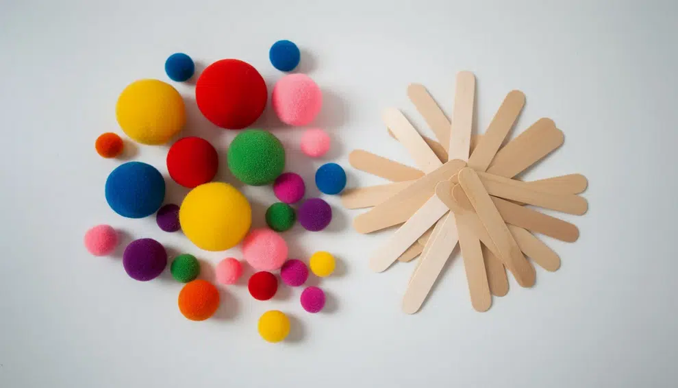 An assortment of colorful pom poms in various sizes is displayed alongside wooden popsicle sticks on a white table, perfect for imaginative play and simple crafts for younger kids. This vibrant collection encourages creativity and fine motor skills as children explore fun shapes and projects.
