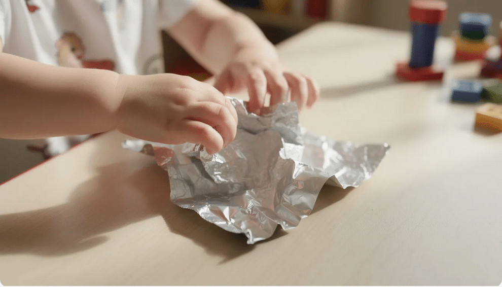 A preschooler's small hands are crumpling a shiny piece of aluminum foil on a table, engaging in a fun art activity that enhances their fine motor skills and hand-eye coordination. This sensory experience allows them to explore textures and shapes, making it a wonderful creative outlet for toddlers.
