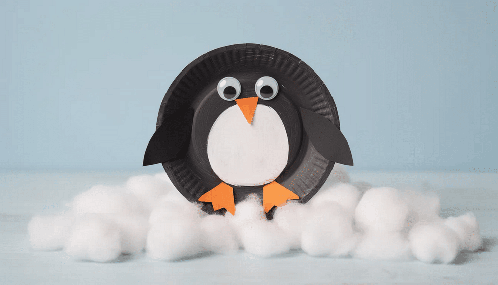 The image features a charming paper plate penguin craft sitting on a base of fluffy cotton ball snow, showcasing a fun winter craft perfect for preschoolers. The penguin, made from black paper and decorated with googly eyes, embodies the playful spirit of winter art projects and is an excellent way to enhance fine motor skills in kids.