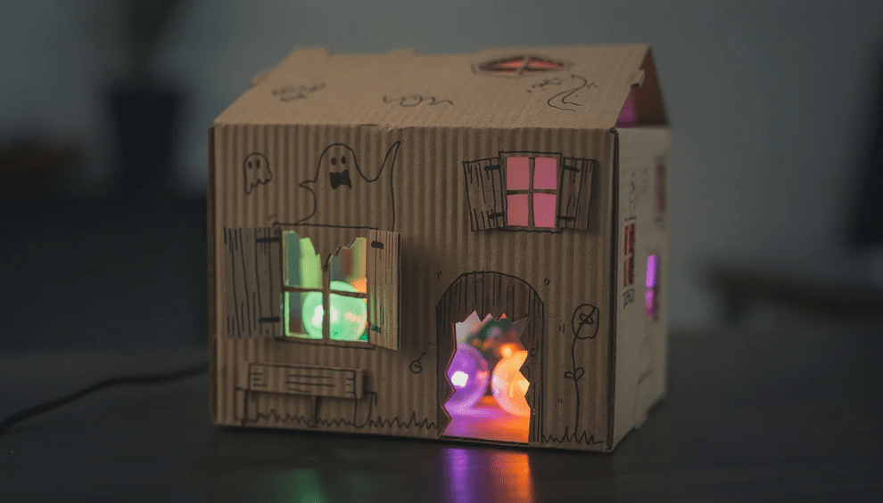 Spooky Cardboard Box The image shows a spooky cardboard box haunted house decorated with colorful construction paper and LED lights glowing through cut-out windows, perfect for Halloween crafts. This fun Halloween design is an easy project that encourages kids to get involved in the spooky season festivities.