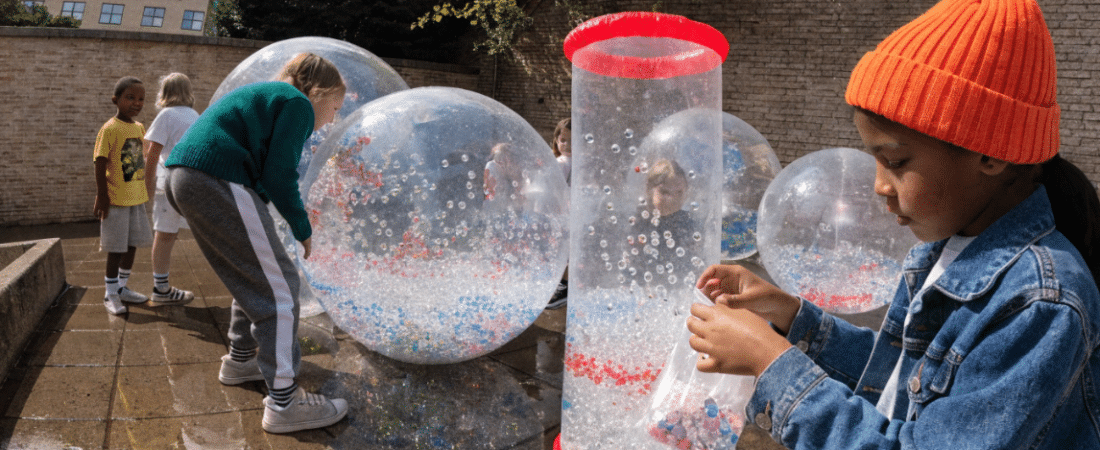 Children play outdoors with large clear inflatable balls filled with colorful confetti and a tall clear tube, enjoying sensory art activities for special needs. One child in a denim jacket and orange hat examines confetti near the tube, while others interact with the balls.