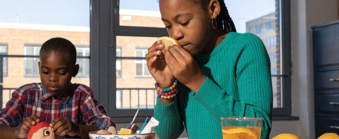Teaching 5 Senses Art and Craft Activities A girl in a green sweater smells a lemon slice at a table, while a boy next to her plays with a toy. There are lemons, cookies, and a glass of lemon water on the table. Sunlight streams through large windows behind them.