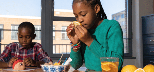 Teaching 5 Senses Art and Craft Activities A girl in a green sweater smells a lemon slice at a table, while a boy next to her plays with a toy. There are lemons, cookies, and a glass of lemon water on the table. Sunlight streams through large windows behind them.