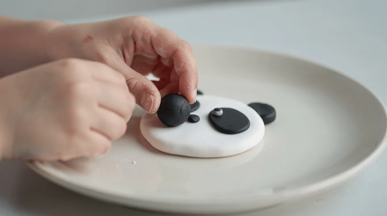 99781e3c-4efa-418e-bd13-48adf9227120 A close-up image shows small hands pressing a tiny black polymer clay ball onto a white circle on a ceramic plate, creating a panda eye. The scene highlights the delicate process of crafting with durable polymer clay, inviting viewers to discover the intricate details of this art.